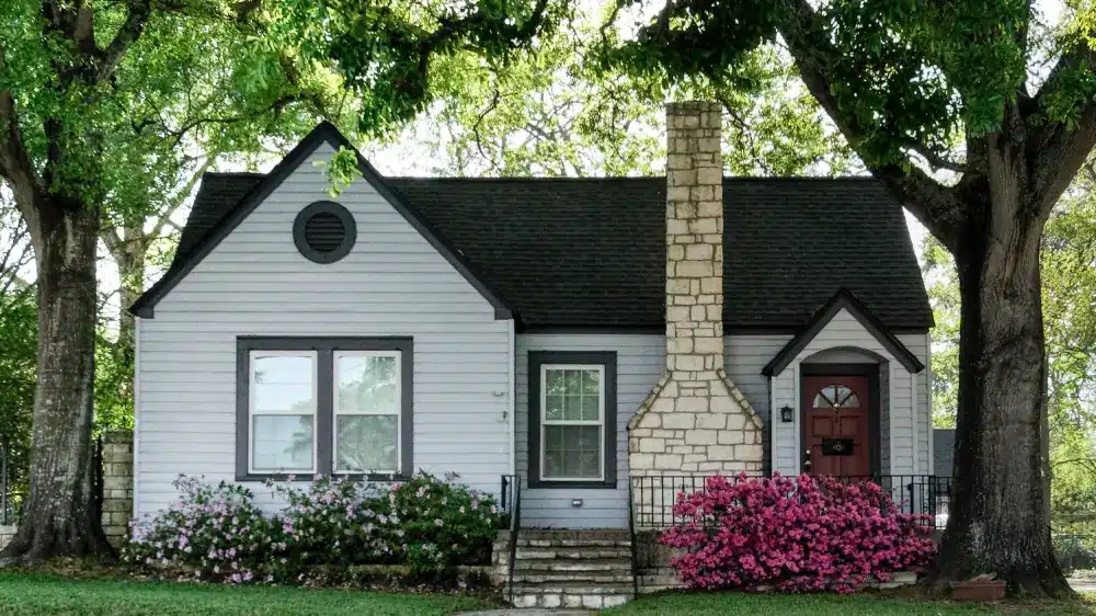 A small gray house with dark roof shingles, prominent stone chimney, and flower bushes in front, surrounded by large trees and green grass.