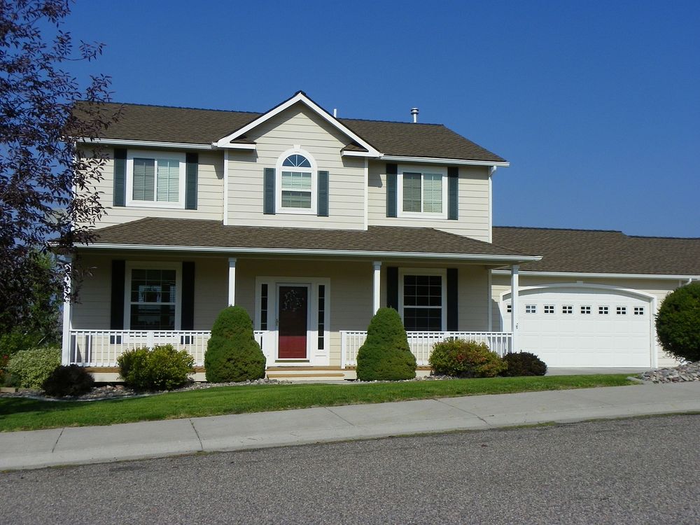 Two-story suburban house with beige siding, white trim, a covered front porch, and an attached two-car garage, viewed from the street on a sunny day.