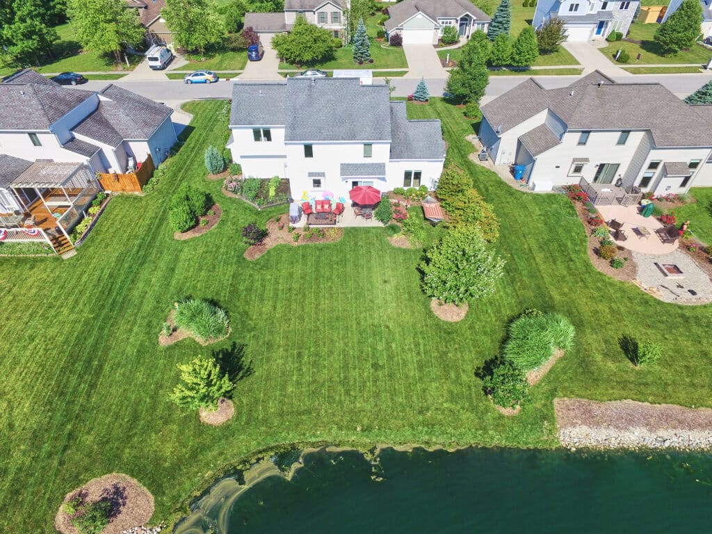 Aerial view of suburban houses in Grey County for home buyers, with landscaped backyards facing a pond, featuring lawns, patio furniture, gardens, and a stone-edged shoreline.