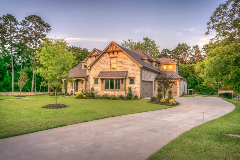 Large brick house with gabled roof, three-car garage, manicured lawn, curved driveway, and trees surrounding the property at sunset.