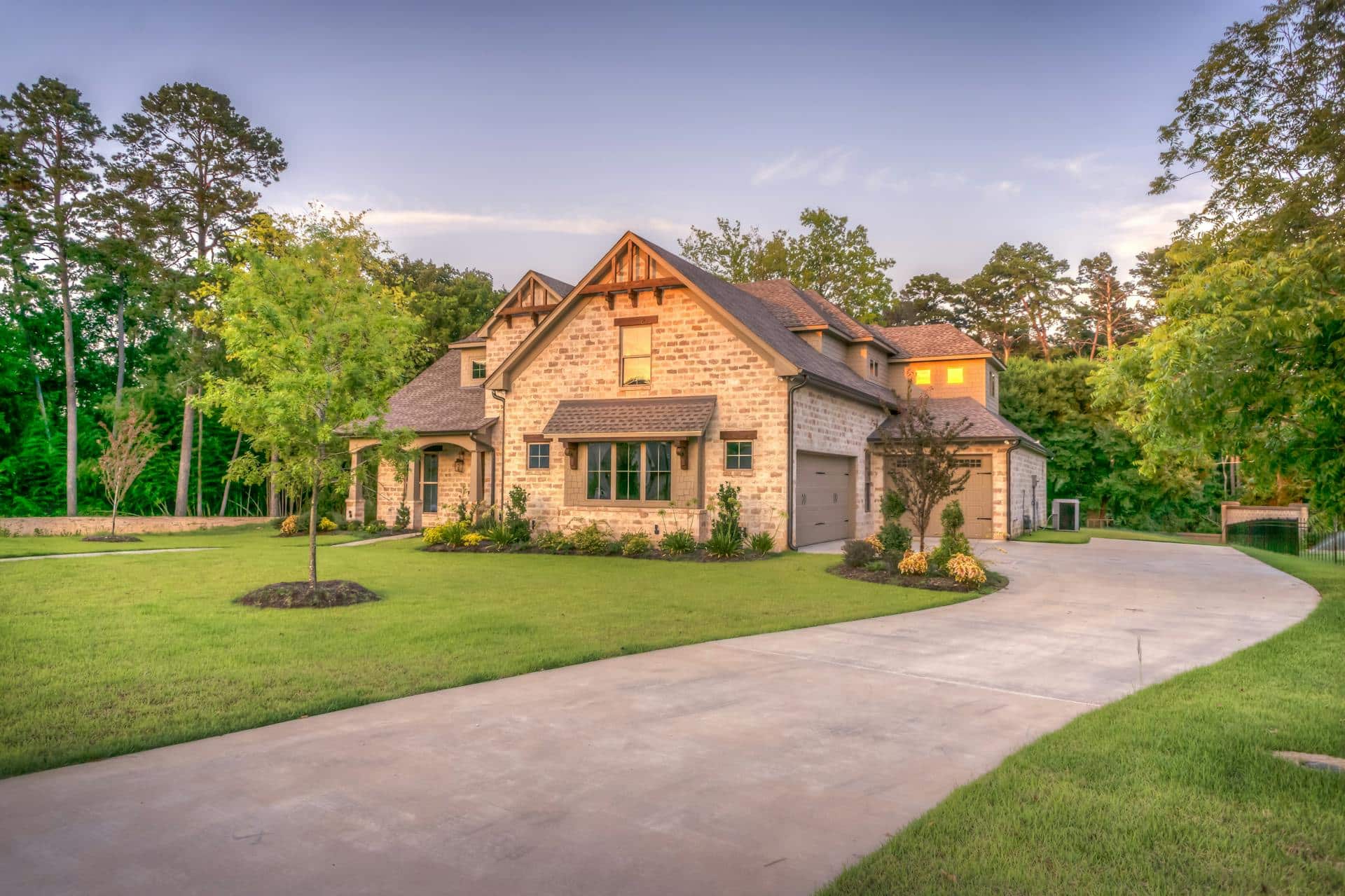Large brick house with gabled roof, three-car garage, manicured lawn, curved driveway, and trees surrounding the property at sunset.