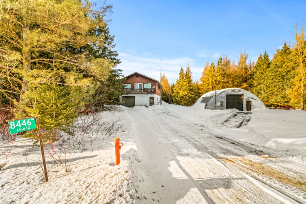 A house with a garage and a metal outbuilding sits amid snow-covered ground and trees; a green address sign reads "8446 Line 12.
