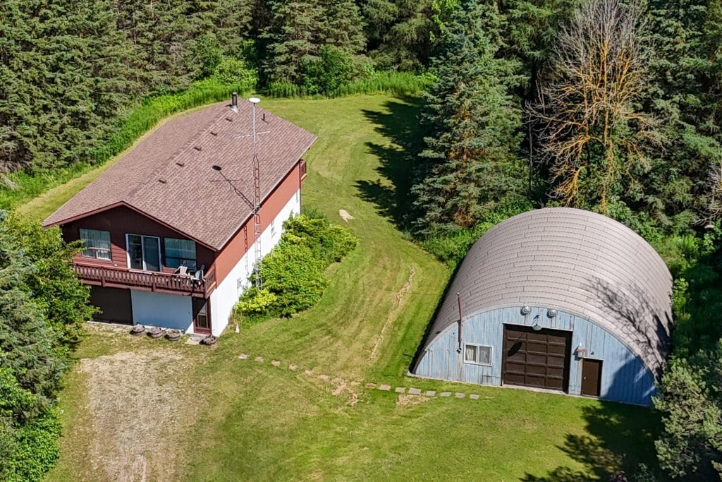 Aerial view of a two-story house with a balcony next to a large Quonset hut garage, surrounded by grass and dense trees.