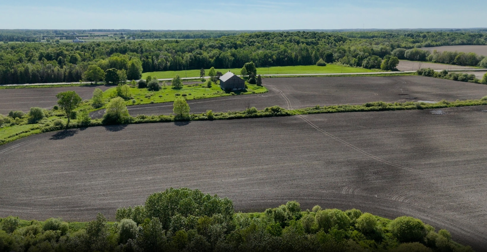 Aerial view of a rural landscape with plowed fields, a small cluster of trees, and a barn surrounded by greenery under a clear sky.