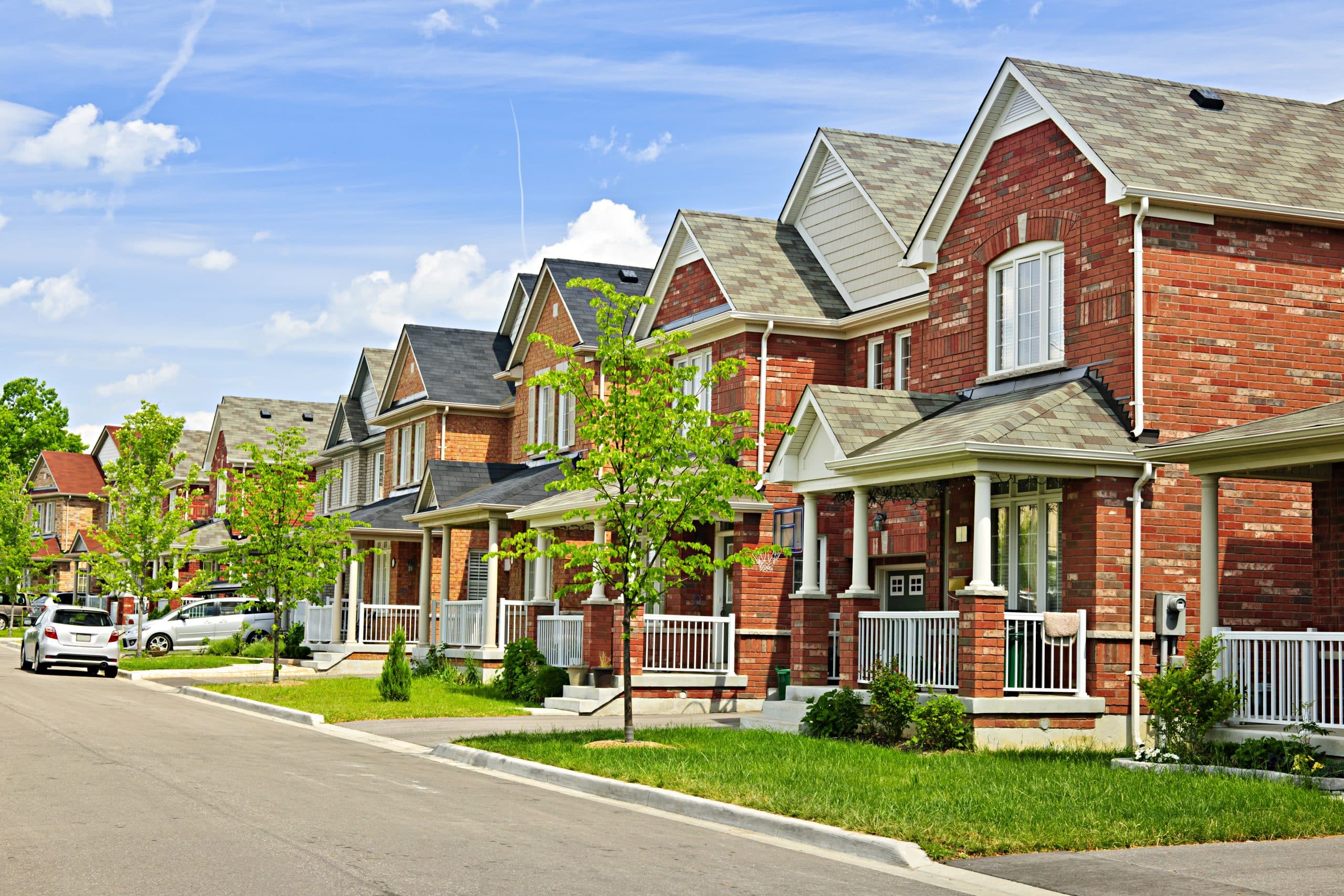 Row of modern suburban brick houses with front porches, small trees, and green lawns on a sunny day. Discover charming homes for sale in Grey County along this quiet residential street with several cars parked nearby.