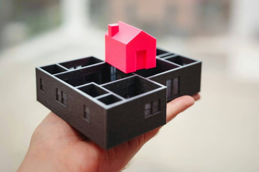 A hand holds a 3D-printed black floor plan model with a red house structure placed on top.