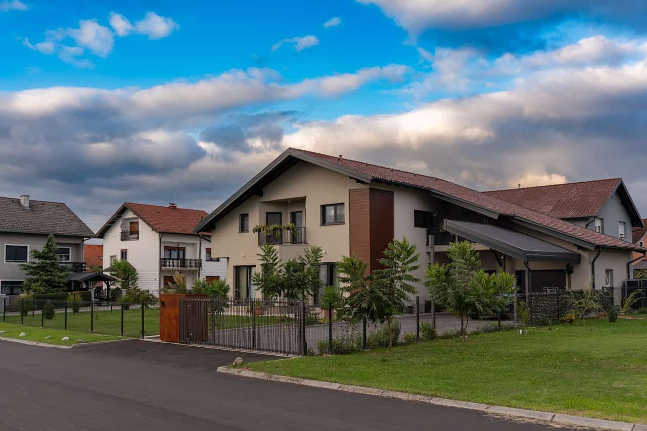 Modern suburban houses with sloped roofs, fenced yards, and well-kept lawns under a partly cloudy sky.
