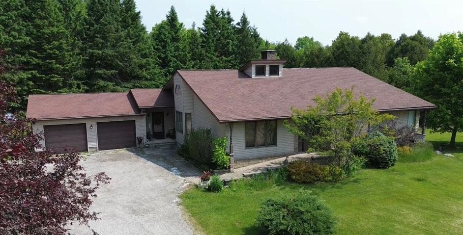 Single-story house with a brown roof, attached garage, and gravel driveway, surrounded by green lawn, trees, and shrubs under a clear sky.
