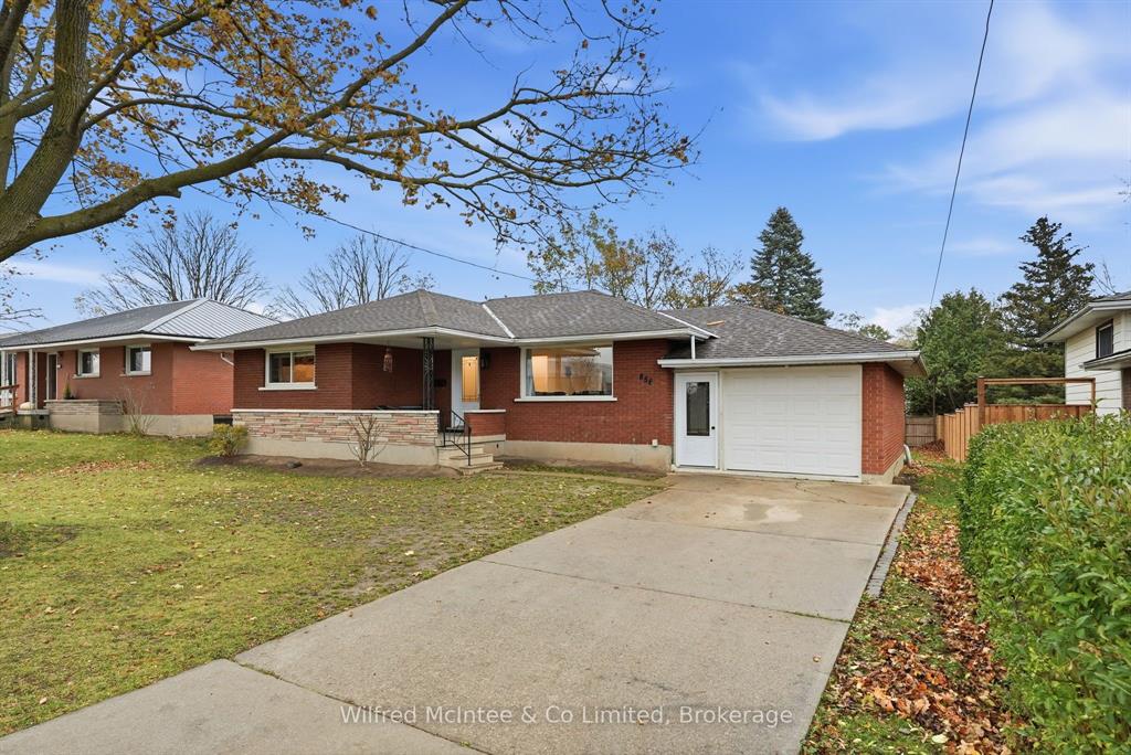 Single-story brick house with attached garage, large driveway, and minimal landscaping on a residential street under a partly cloudy sky.