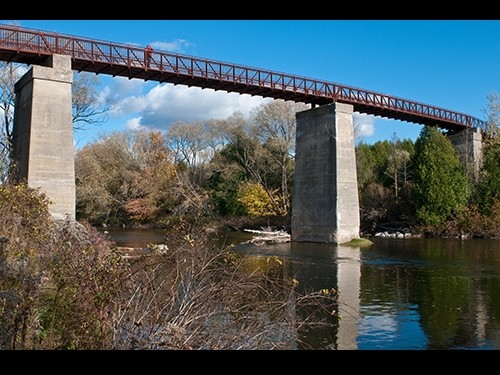 A tall pedestrian bridge with concrete supports spans over a river, surrounded by trees and vegetation under a blue sky with clouds—a scenic highlight of Hanover Ontario real estate.