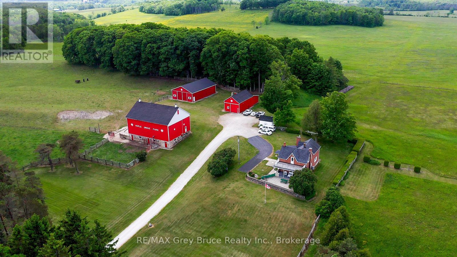 Aerial view of a rural property with a red barn, two red outbuildings, a house, driveway, and several vehicles, surrounded by green fields and trees—perfect for those searching for farms for sale in Grey County.