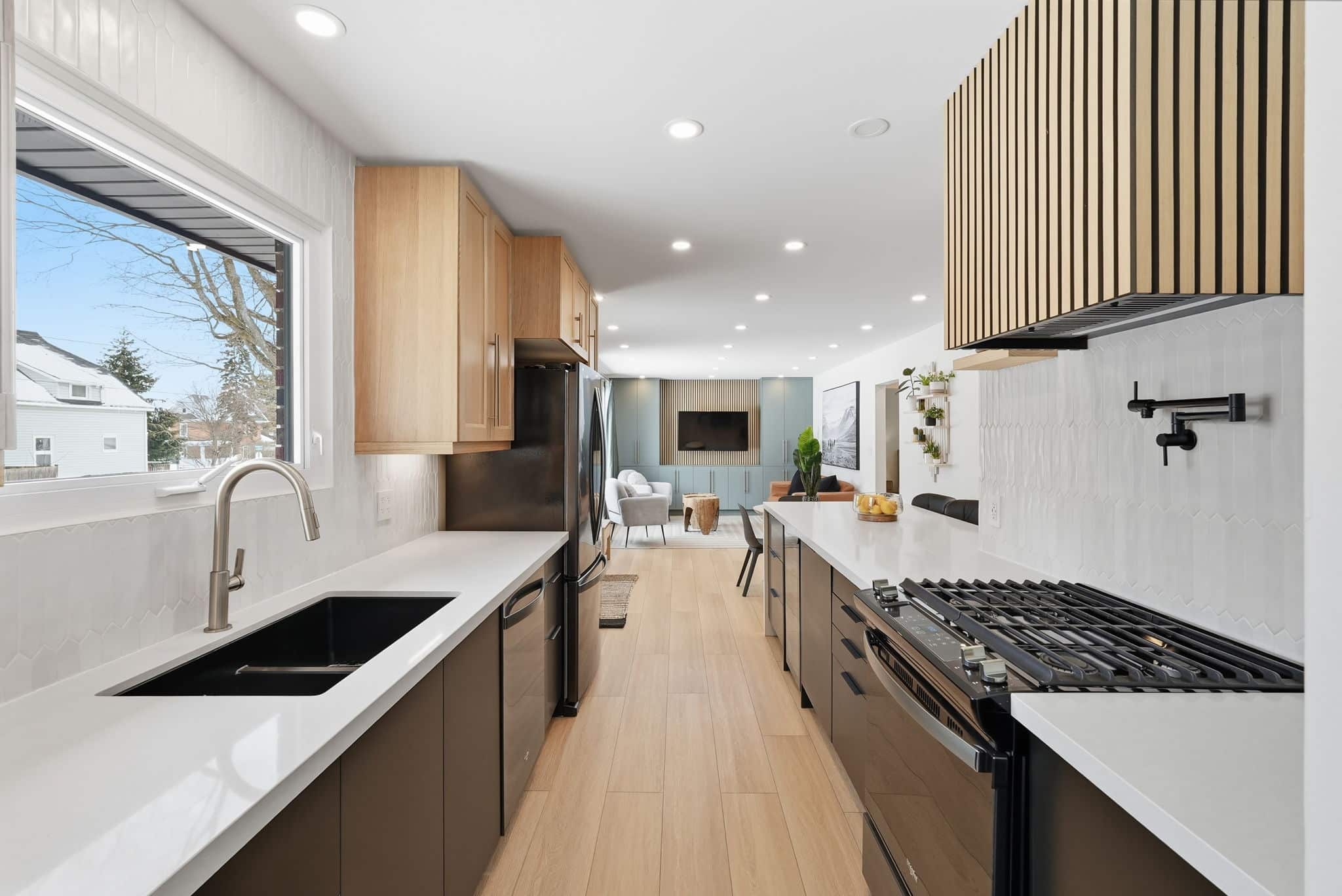 Modern kitchen with black appliances, wood cabinetry, white countertops, gas cooktop, and a large sink under a window; open to a bright living area.