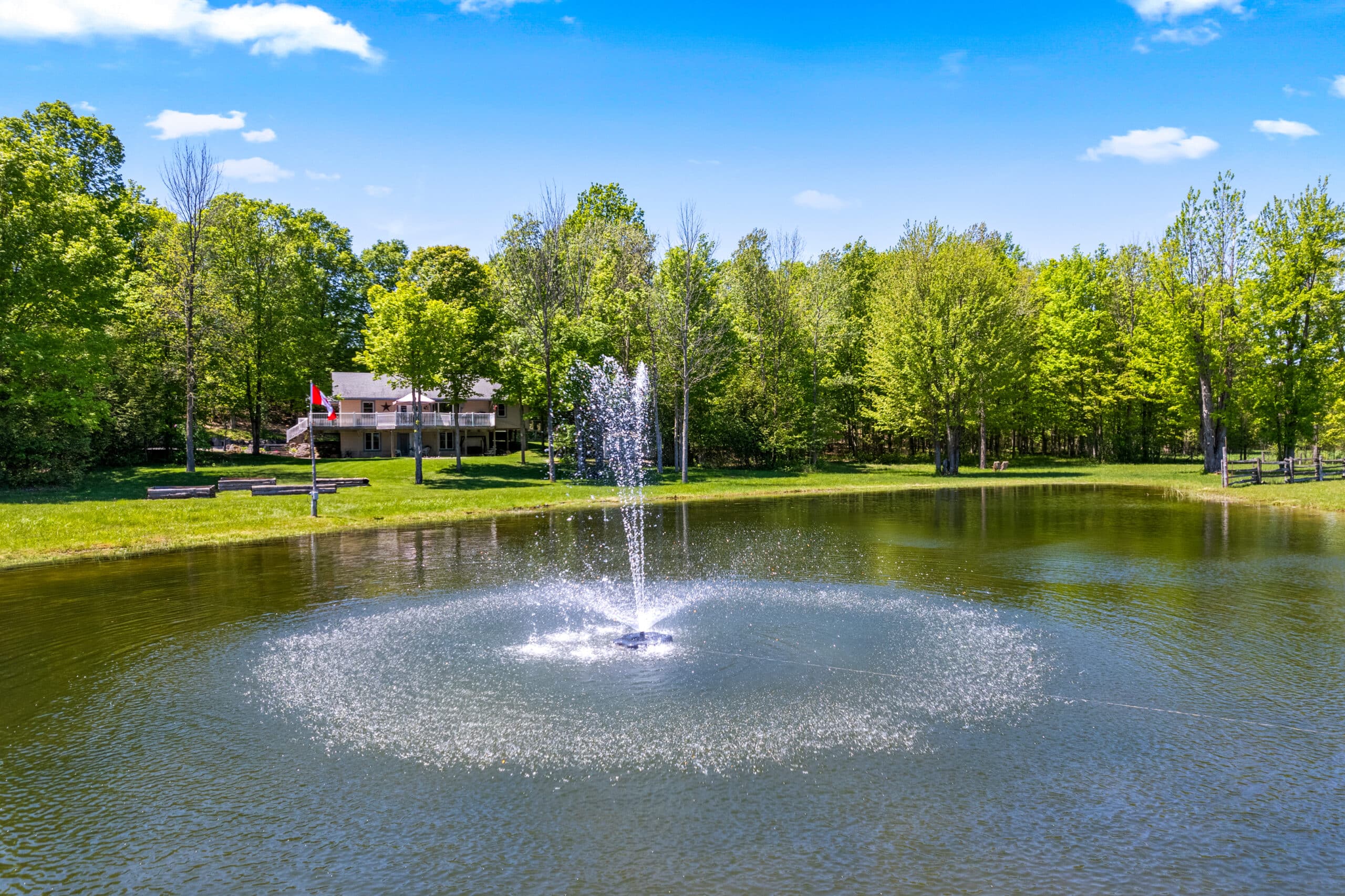 A water fountain sprays in the center of a pond surrounded by green trees, with a house and Canadian flag visible in the background.