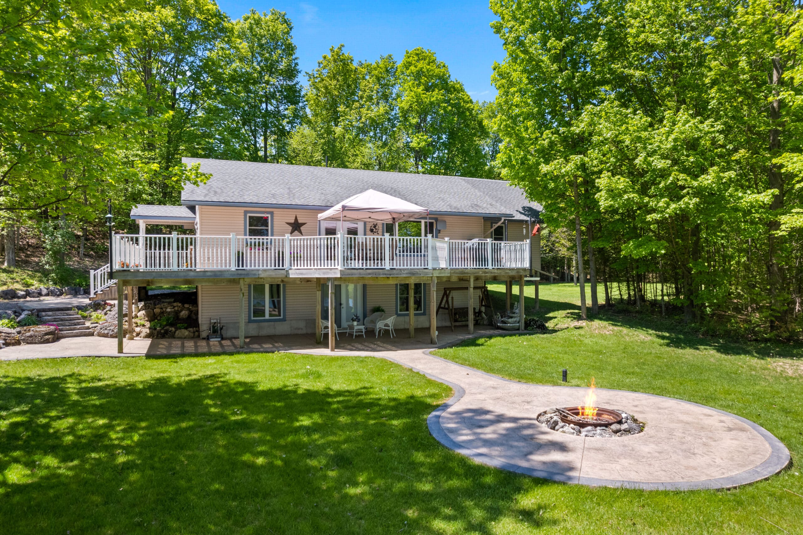 A two-story house with a large elevated deck, shaded seating area, and a circular fire pit on a concrete patio surrounded by green lawn and trees.