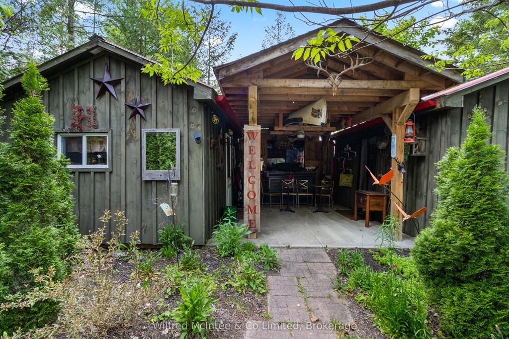 Rustic wooden shed with a covered open-air workspace, "WELCOME" sign, stars on the exterior, surrounded by greenery and trees.