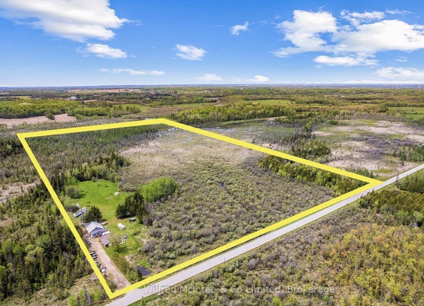 Aerial view of a large rectangular rural property outlined in yellow, featuring a house, trees, and open fields, bordered by a road and surrounded by forested land.