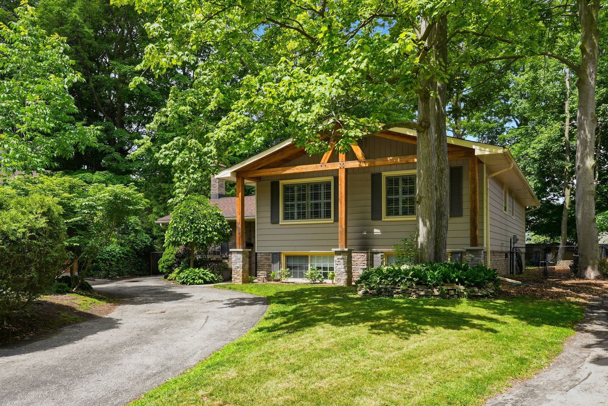 A split-level house with brown siding and a covered porch, surrounded by trees and greenery, with a curved driveway and a well-kept lawn.