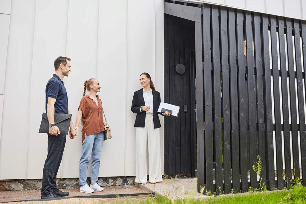 A real estate agent stands outside a modern house, showing the property to a couple holding hands.