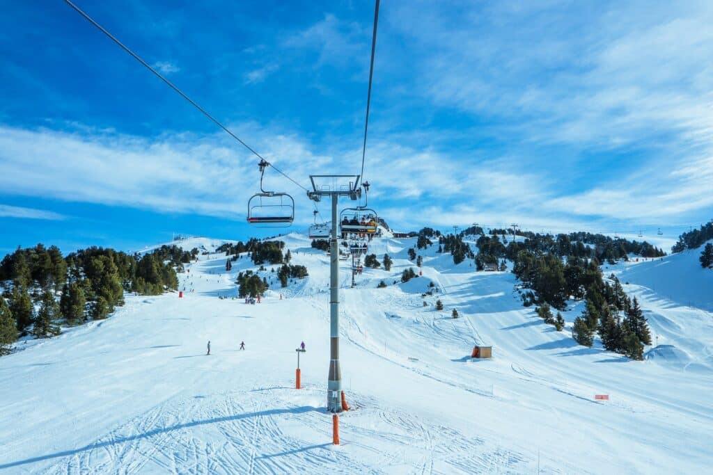 A ski lift ascends over a snowy mountain slope with scattered trees, skiers below, and a blue sky overhead.