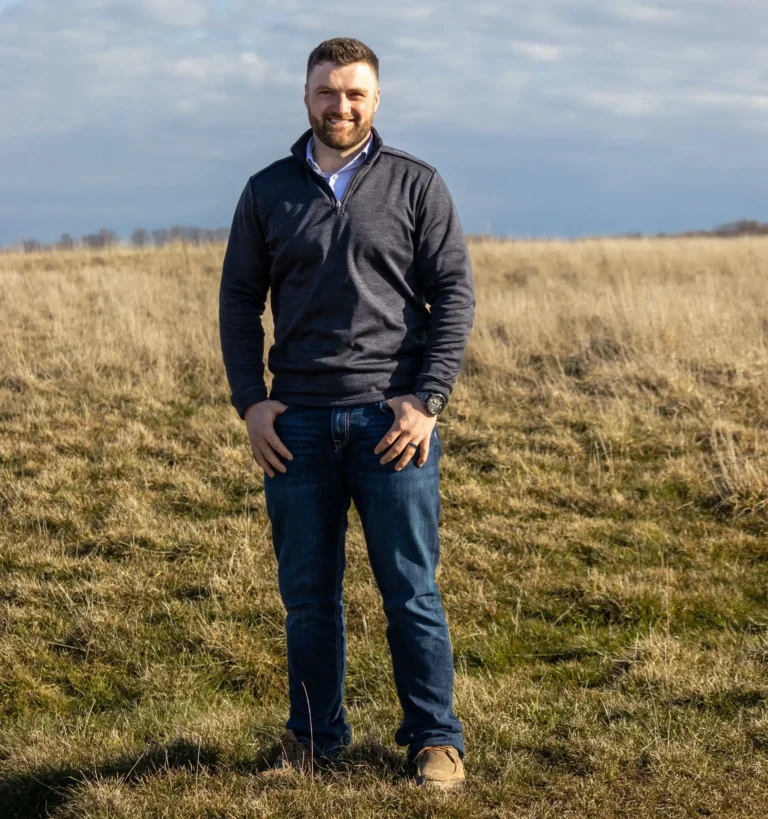 A man wearing a dark sweater and jeans stands smiling in an open grassy field under a partly cloudy sky.