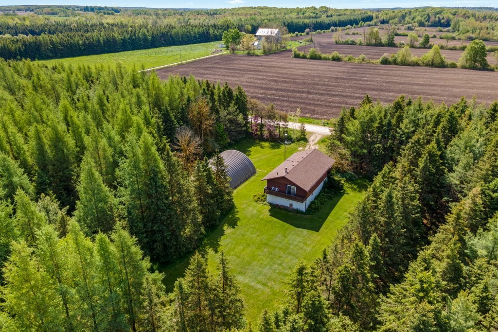 Aerial view of a house and a metal shed surrounded by dense trees, with open fields and a distant farmhouse in the background.