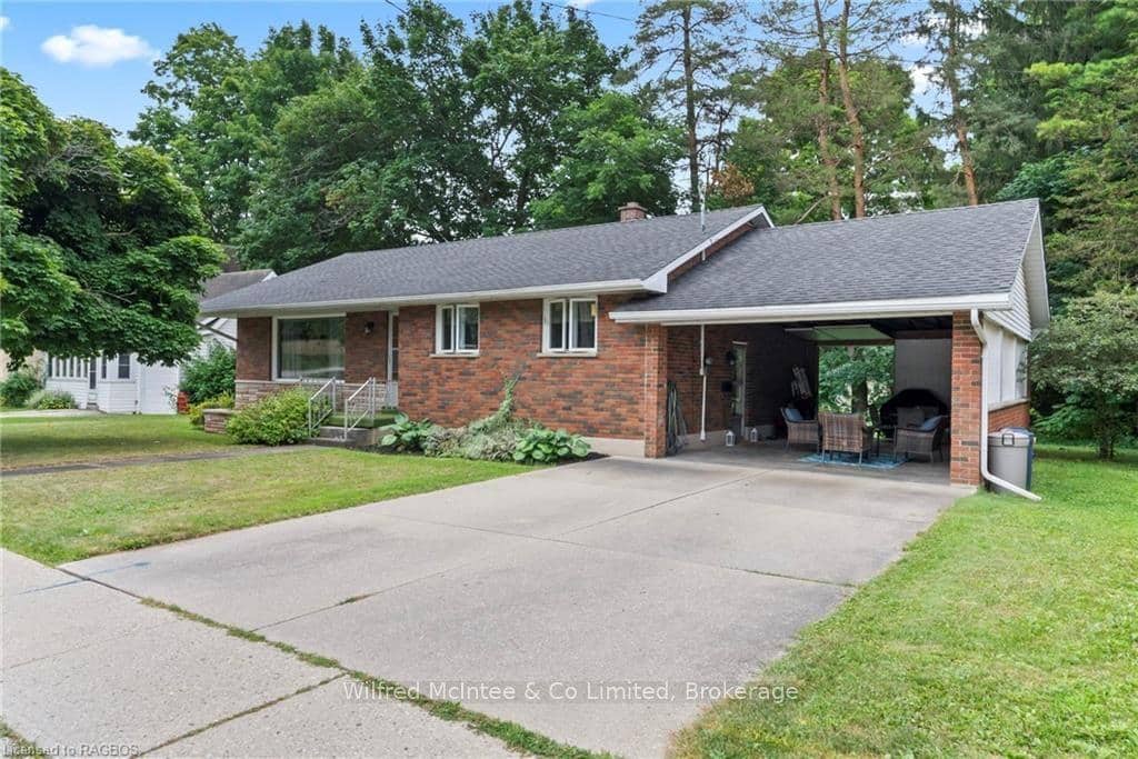 Single-story brick house with a sloped roof, attached carport, paved driveway, and surrounded by trees and greenery.