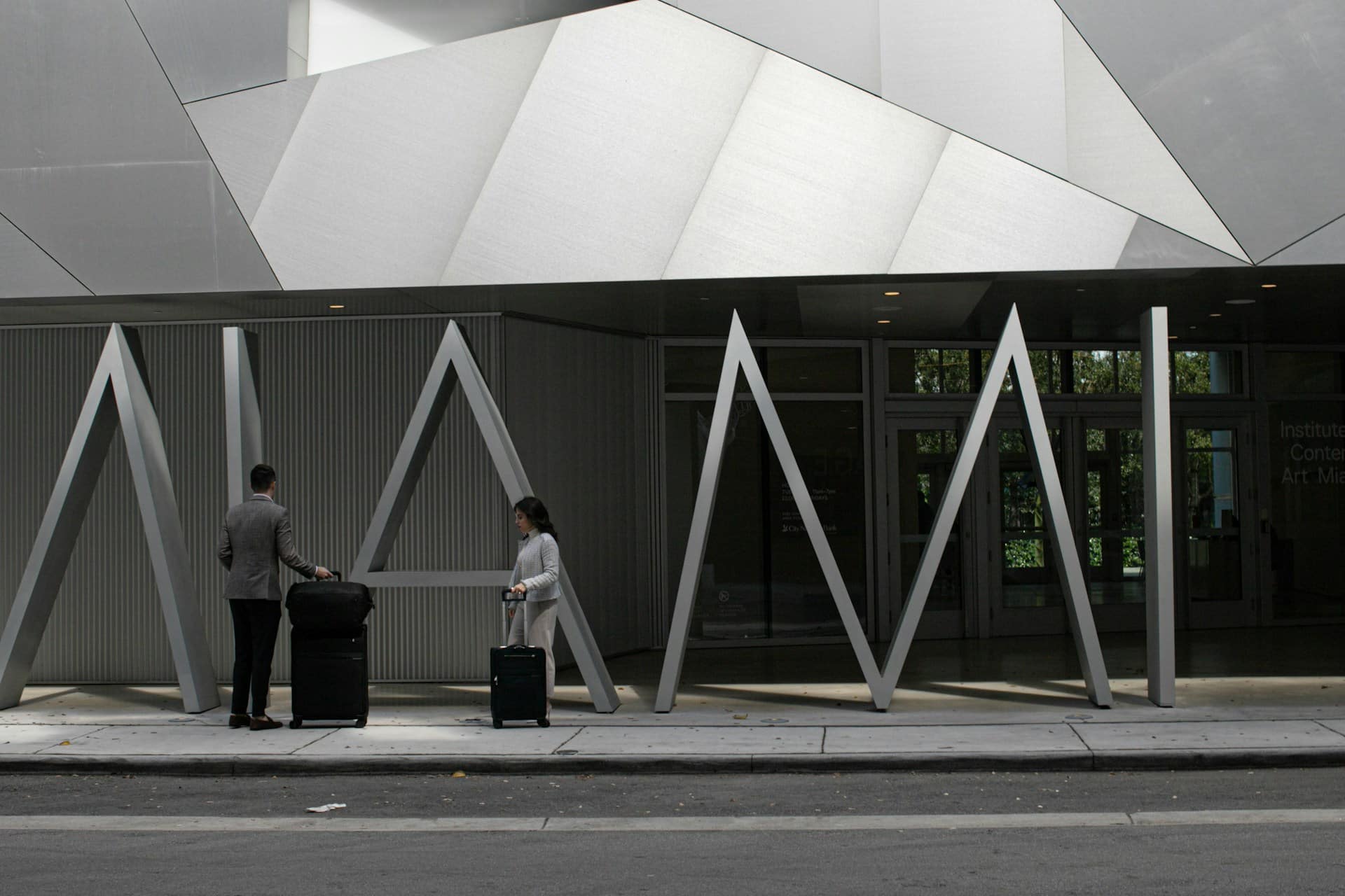 A man and a woman with suitcases stand outside a modern building with geometric metal architecture and large angular columns.