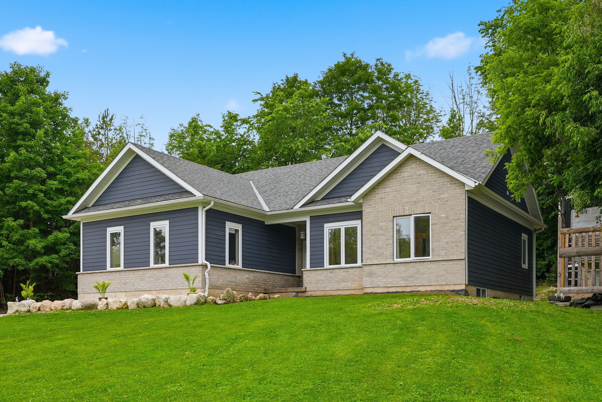 Single-story house with blue siding and light stone accents, large windows, gable roof, and a well-maintained lawn, surrounded by green trees under a clear sky.